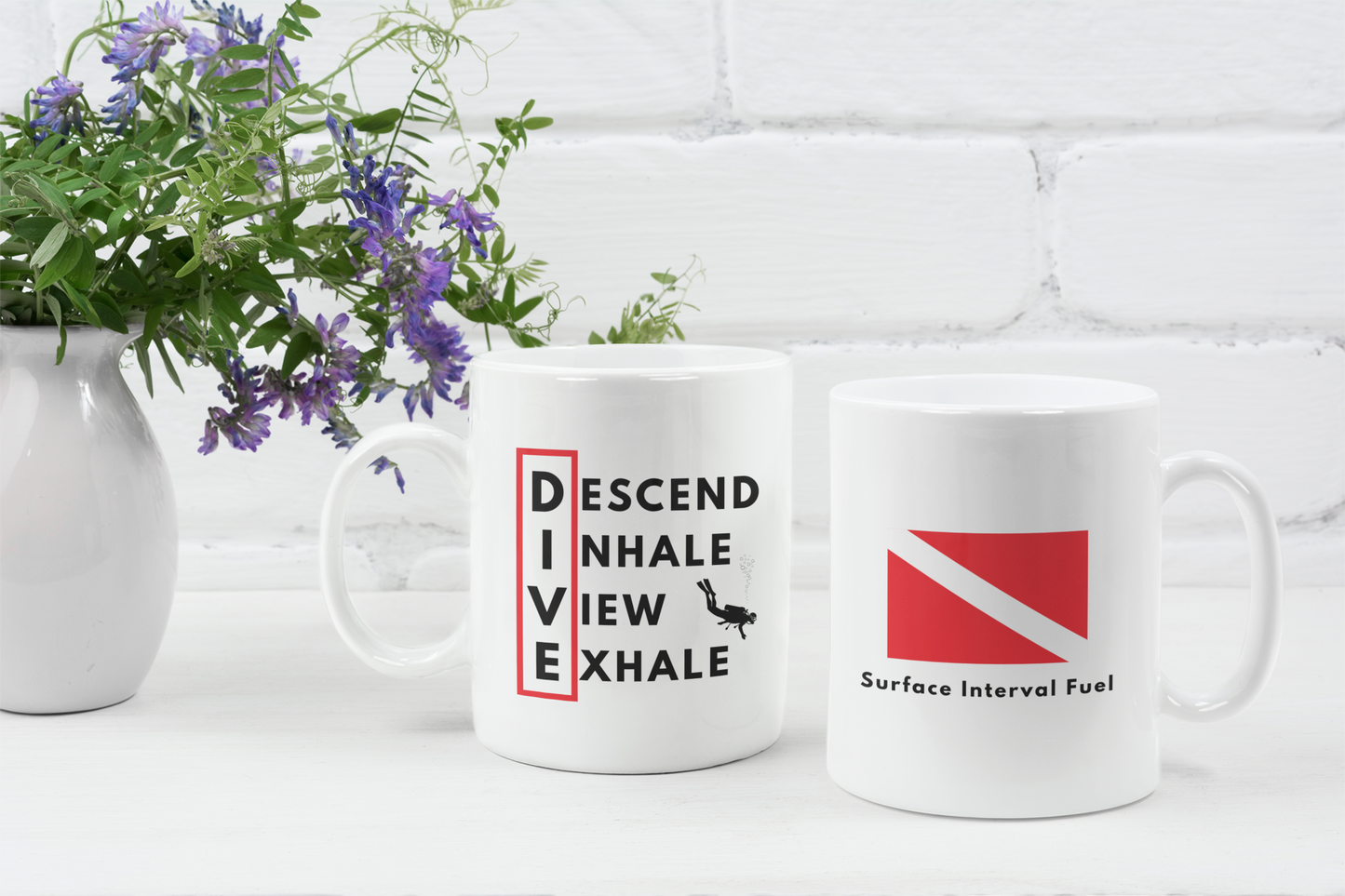 Two mugs with motivational and diving-themed designs on a white surface with a floral arrangement.