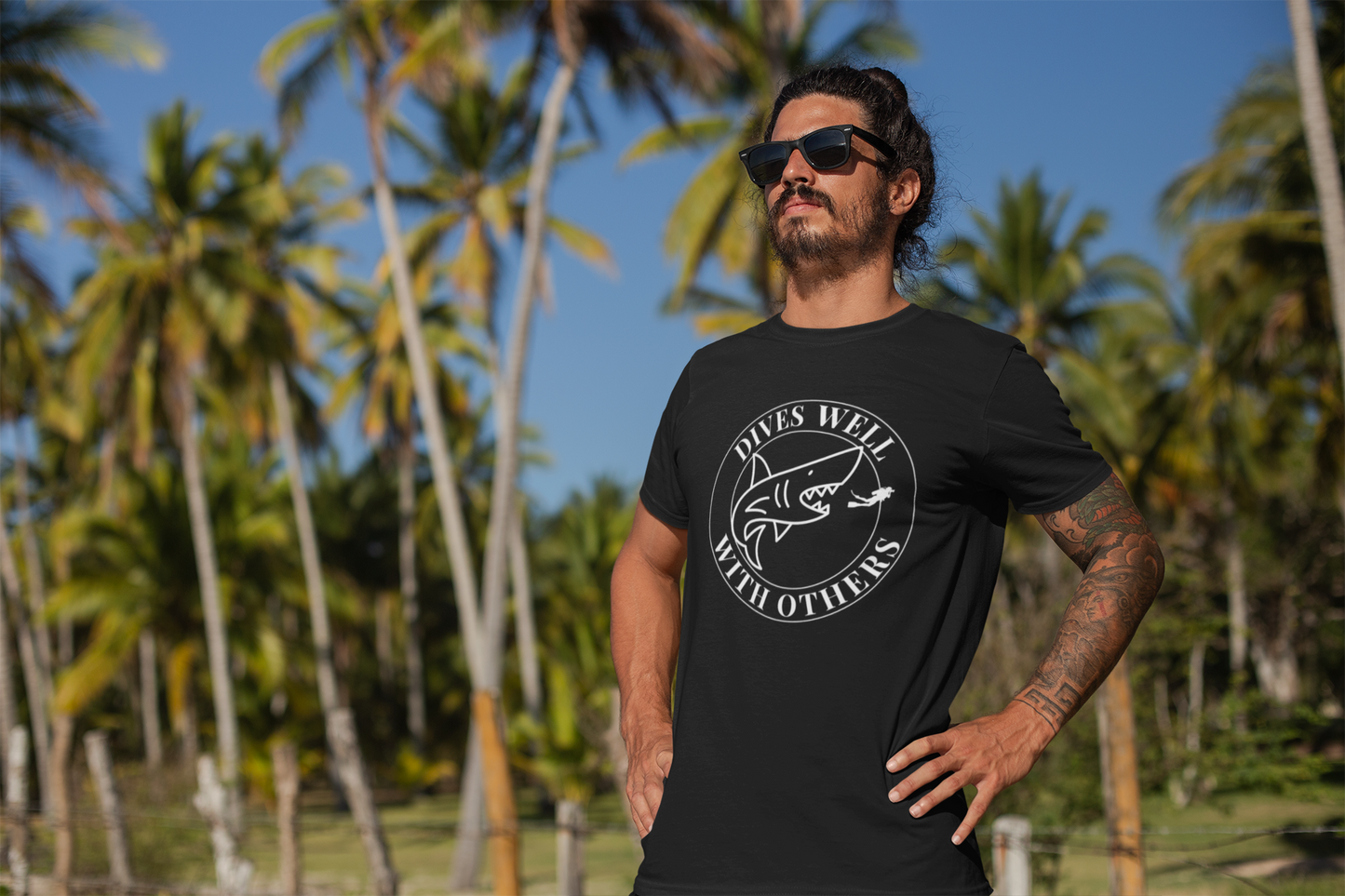 Man wearing a black t-shirt with a graphic design and text, standing in front of palm trees.