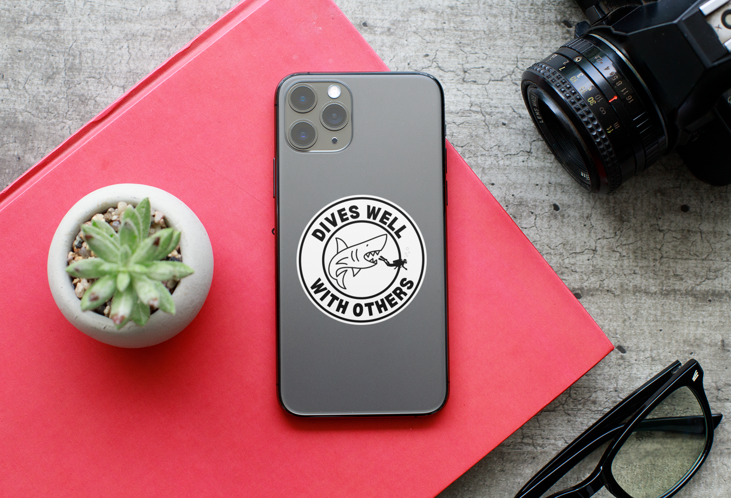Phone case with 'Dives Well With Others' design on a red surface with camera and glasses. Scuba Diver.