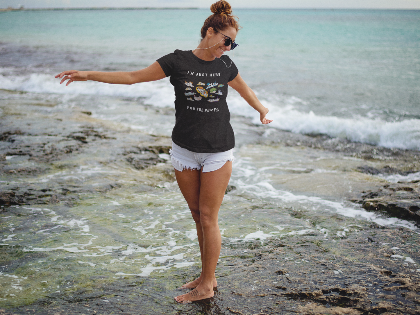 Woman standing on a rocky beach wearing a black t-shirt with nudibranch images and white shorts.
